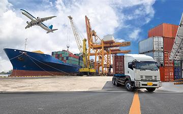 Truck parked on a dock next to cargo containers and a cargo ship with an airplane flying above