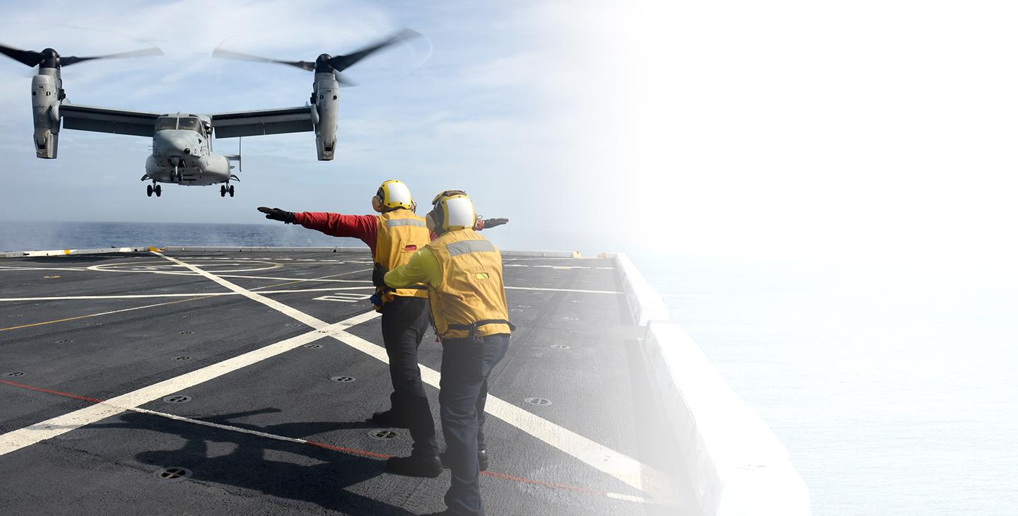 Two people using military satcom to communicate with a MV-22 landing on an aircraft carrier at sea